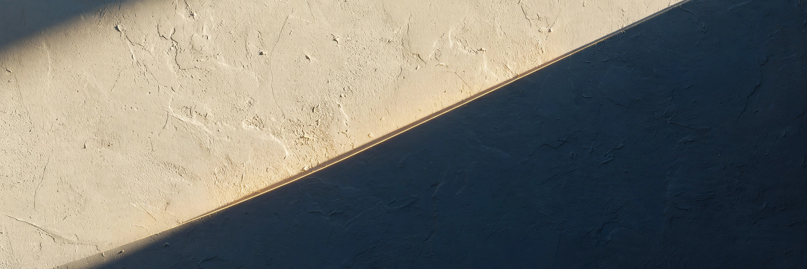 Warm morning light raking across a textured plaster wall, a deep navy shadow cut by a thin line of golden light